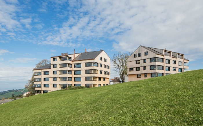 Two multi-storey timber residential buildings on a green meadow against a blue sky